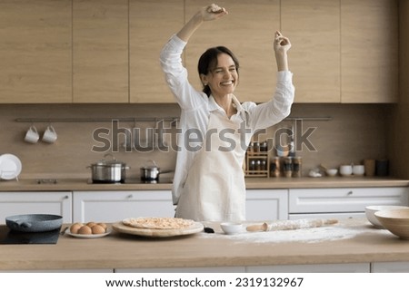 Happy young attractive woman dancing in kitchen while cooking homemade pizza, female in apron moving to favourite music, feels carefree, enjoy pastries preparation on weekend at home. Culinary, hobby Royalty-Free Stock Photo #2319132967