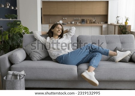 Cheerful millennial woman relax on sofa in studio apartment, leaned on soft comfortable couch with hands behind head, smile enjoy carefree leisure at smart home with climate control for comfort life Royalty-Free Stock Photo #2319132995
