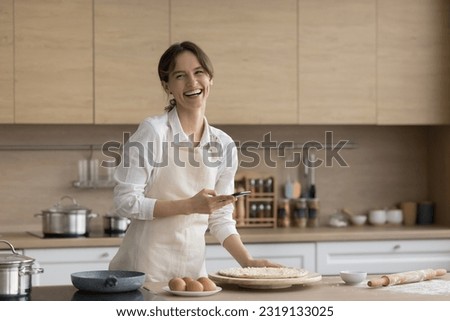 Laughing woman in apron holds smart phone, making pictures of fresh homemade pizza. Female food blogger share Italian cuisine recipe with channel followers, enjoy cookery process in modern kitchen