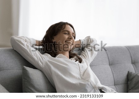 Attractive young 30s female breath fresh conditioned air, relaxing leaned on cozy couch looks carefree, smile, feels happy, enjoy stress-free weekend leisure at modern smart home with climate control Royalty-Free Stock Photo #2319133109