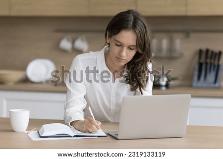 Attractive focused young woman takes notes, sitting at kitchen table with laptop, working or studying on-line at home, e-learning, planning workday tasks in personal organizer, writing things to-do