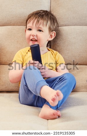 The joyful child is seated on the sofa holding the TV remote, watching cartoons with a beaming smile. A little baby is lying on the couch, watching a cartoon on the television set with happiness.
