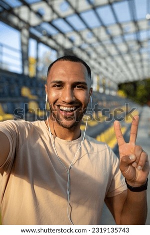Close-up photo. Vertical photo. Young African American male sportsman stands in headphones at the stadium, takes a selfie on the phone, talks on a video call. Shows a super gesture to the camera. Royalty-Free Stock Photo #2319135743