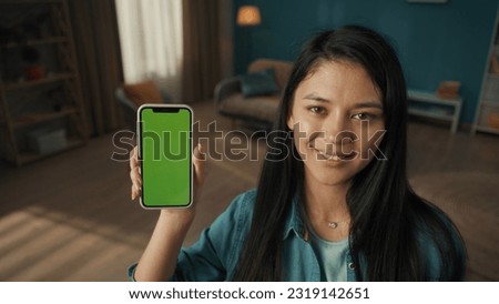 Young smiling woman shows smartphone with green screen. Portrait of an Asian woman with a smartphone in the living room closeup. Technology, gadget, connection. Advertising area, workspace mock up.