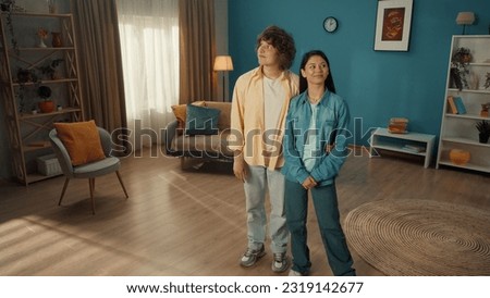 A young couple is standing in a cozy living room and examining it. The curlyhaired young man put his arm around the girl's waist. Young people admire their family nest. Royalty-Free Stock Photo #2319142677