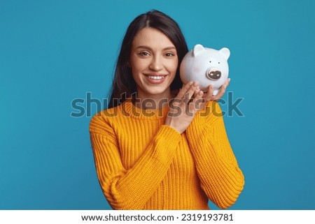 Cheerful young woman in knitted orange sweater holding white piggy bank with lots of money near face isolated over blue background