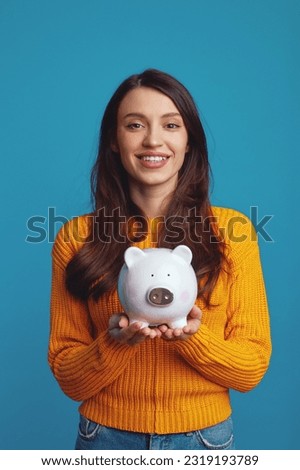 Vertical photo of happy young woman in casual orange sweater holding white piggy bank with lots of money isolated over blue background