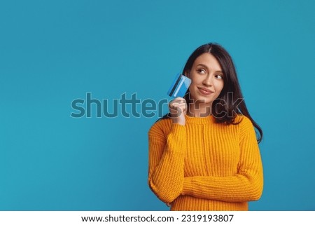Thoughtful young girl wearing yellow clothes holding plastic credit card near face and looking at free empty space, isolated over blue background 