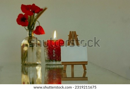 A wooden miniature easel stands on a table next to a glass vase with red poppies and a red candle with a burning flame. Decor for cozy home. Drawing Lessons, art and hobby concept. Draw a picture.