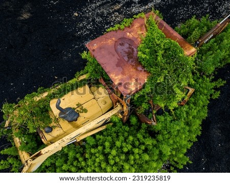 A low altitude, aerial view over an old and rusted tractor, taken over by nature with green vines and bushes. It is surrounded by black mulch and shot on a sunny morning.