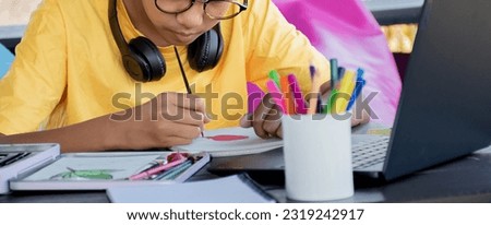 Asian boy in yellow shirt spending his free time with drawing, sketching, coloring and painting various shapes of animals and fruits on table in his house, recreational activity concept,cropped shot.