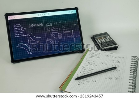 Tablet with math problems displayed. A notebook with notes, a calculator and a pen.

