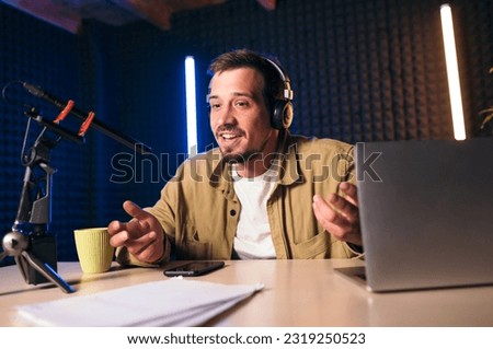 Young stylish man in mustard shirt with headphones gesturing at microphone and sharing story with audience while sitting at desk in studio with neon lighting and recording podcast Royalty-Free Stock Photo #2319250523