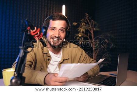 Smiling radio host with headphones reading news from paper into studio microphone at radio station with neon lights. looking at camera