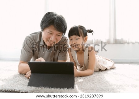 Portrait of Asian father and cute little daughter watching cartoons using digital tablet gadget e-books for kids on-line lying on carpet at home. Parental control protect from bad content concept