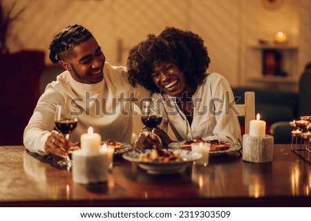 Portrait of a cheerful happy african american couple drinking wine and having romantic dinner at home. Home date night with home cooked meal and red wine. Candle light and moody atmosphere. Copy space Royalty-Free Stock Photo #2319303509