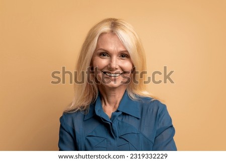 Portrait of cheerful happy beautiful elderly blonde woman 60s in nice blouse shirt posing on beige studio background, smiling at camera. Seniors lifestyle, older adults concept
