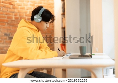 Online Lecture. Side View Of Korean Student Guy With Headset Using Laptop Computer And Writing Notes, Sitting At Table At Home. Teen Engaging In Remote Education Doing Homework At Portable PC Royalty-Free Stock Photo #2319332255