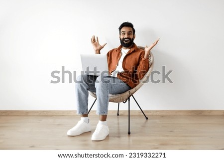 Online Luck. Glad arab guy sitting in armchair with laptop and raising hands in joy, receiving great news and celebrating success over white wall background. Wow internet offer