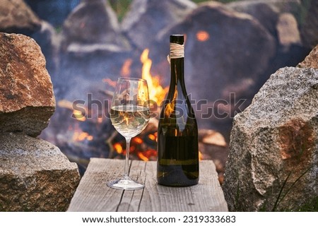 Romantic picture of the glass of white wine and rustic style bottle from small family winery in front of burning stone campfire during the summer sunset in the south moravian region of Czech Republic.