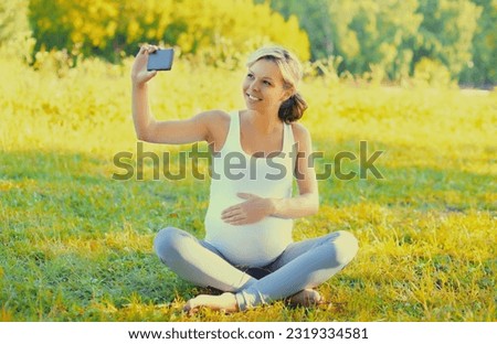 Happy smiling young pregnant woman taking selfie with smartphone sitting on the grass in summer park