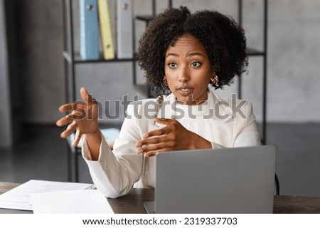 Confident corporate beautiful young african american businesswoman in white formal outwear sitting at desk in front of laptop, talking and gesturing, have interview at office, copy space Royalty-Free Stock Photo #2319337703