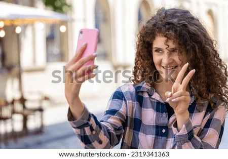 Pretty young woman blogger taking selfie on smartphone, communicating video call online with subscribers, recording stories for social media vlog outdoors. Girl walking in urban sunshine city street