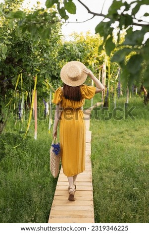 Woman wearing a straw hat walking along the path in the summer garden back to the camera. Female enjoing spring evening in the park. Girl in the yellow dress with a wicker bag and flower bouquet. Royalty-Free Stock Photo #2319346225