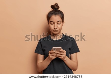 Photo of serious dark haired latin woman concentrated in smartphone chatting on internet working or studying online watches video dressed in casual black t shirt isolated over brown background