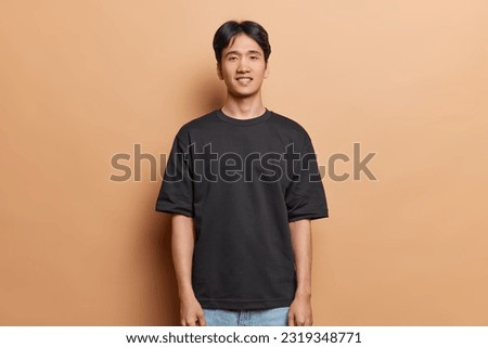 Handsome cheerful Chinese guy with pleasant smile looks directly at camera wears casual black t shirt and jeans keeps arms down isolated over brown background. People ethnicity emotions concept