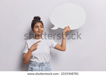 Place your promotional content here. Pleased dark haired Indian woman points at blank speech bubble wears casual t shirt and jeans isolated over white background suggests to write your ideas here Royalty-Free Stock Photo #2319348781