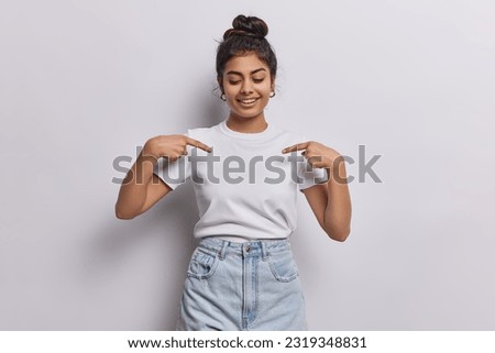 Young dark haired woman captures attention with her vibrant energy with contagious smile directs focus to her blank white tshirt using two fingers shows place for your logo or design poses indoor