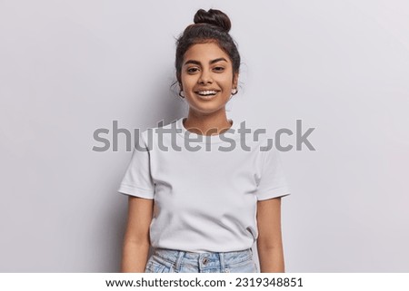 Horizontal shot of Indian woman with dark hair gathered in bun smiles pleasantly being in good mood dressed in casual t shirt and jeans isolated over white background. People and emotions concept