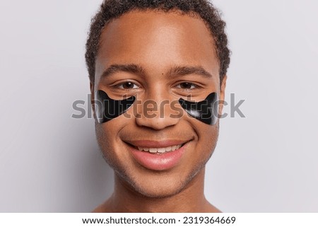 Close up portrait of young man indulges in male beauty care sporting undereye hydrogel black patches on his face to reduce puffiness smiles pleasantly isolated over white background. Personal grooming Royalty-Free Stock Photo #2319364669