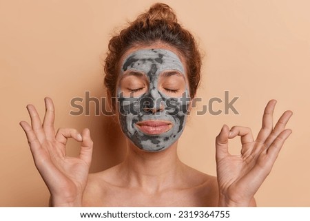 Peaceful woman keeps eyes closed and hands spread wide in zen gesture as she luxuriates in tranquil ambiance applies nourishing beauty mask isolated over beige background. Skin care concept.