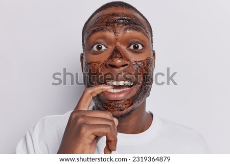 Portrait of dark skinned man applies coffee mask to cleanse his skin aiming for refreshed and healthy appearance bites finger dressed in casual t shirt isolated over white background. Beauty concept
