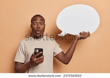 Waist up shot dark skinned adult man wears casual tshirt holds speech bubble and modern smartphone capturing essence of peoples engagement in online conversations isolated over brown background