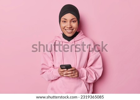 Pleased young Muslim woman using phone application edit pics with funny filters smiles positively wears black hijab and sweatshirt isolated over pink background. People and technology concept