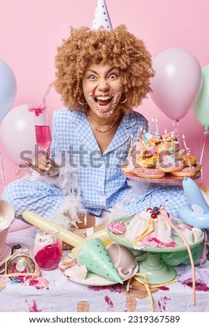Irritated curly haired European woman yells loudly has cream around mouth drinks cocktail and eats tasty creamy desserts isolated over pink background. People partying and celebration concept