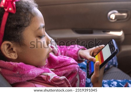 Little Multiracial Girl Captivated by Rainbow Flag on Smartphone in Car Ride. LGTBQ, Diversity, Copy space.