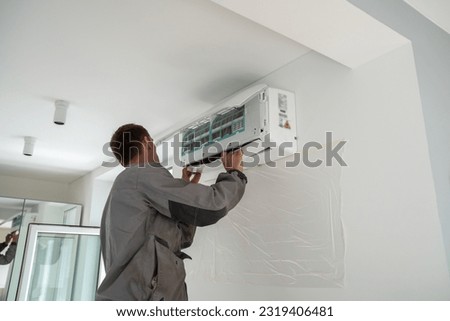 Male worker wearing uniform installing air conditioner in apartment during summer season, man technician standing indoors repairing HVAC system, checking and replacing AC filter Royalty-Free Stock Photo #2319406481