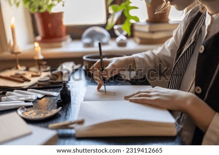 Close up of female artist drawing with ink using dip pen, sitting at table with art supplies in cozy studio, making art, painting process. Creativity and inspiration concept Royalty-Free Stock Photo #2319412665