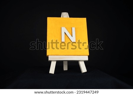 N wooden capital letter and orange blank painting canvas resting on a miniature artists easel isolated on a black background