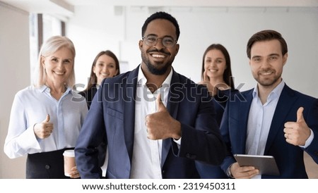 Happy African American business leader and team making thumb up like gesture at camera. Head shot portrait of young millennial businessman, boss standing in front of employees group. Team work concept