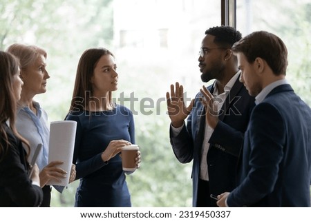 Diverse business team standing, talking at office window, brainstorming on project at casual meeting. African American group leader speaking to employees on briefing. Interns discussing training exam Royalty-Free Stock Photo #2319450337
