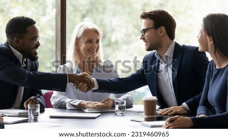 Happy diverse business partners shaking hands over table on partnership meeting, closing deal. Team leader expressing recognition to employee on good work result, achieve, hiring, promotion