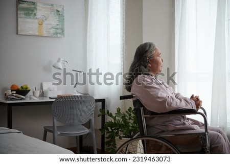 Sick senior woman sitting in wheelchair and looking through window Royalty-Free Stock Photo #2319463073