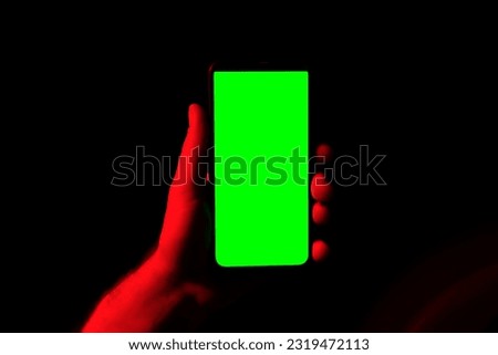 Mobile phone in young man hand. Use smartphone with green screen on red blue neon lighting, dark background. Male showing black phone chroma key display. Close up. Hand lifts device up holds it still.
