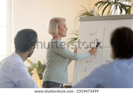 Middle aged businesswoman teacher training staff, pointing at diagram on white board, explaining project strategy to diverse colleagues, mature mentor coach making flip chart presentation Royalty-Free Stock Photo #2319473499