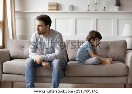 Unhappy young Caucasian man father and little 6s son sit separate ignore each other after family fight. Upset stubborn dad and small boy child avoid talking having quarrel. Generation gap concept. Royalty-Free Stock Photo #2319493141